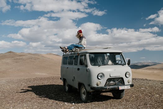 Full length of anonymous female traveler sitting on roof of van in desert under cloudy sky